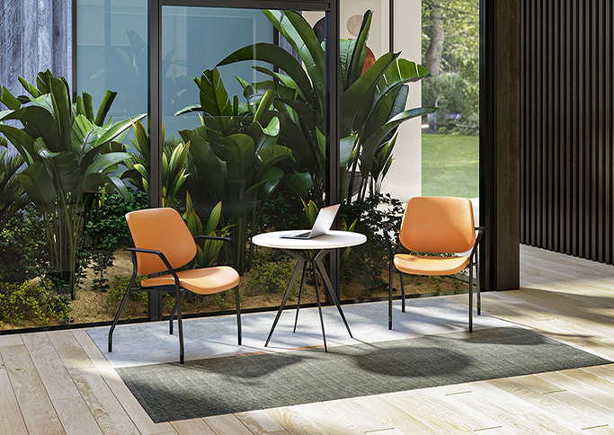 A modern seating area with two orange chairs and a small white table, surrounded by lush greenery and large windows.